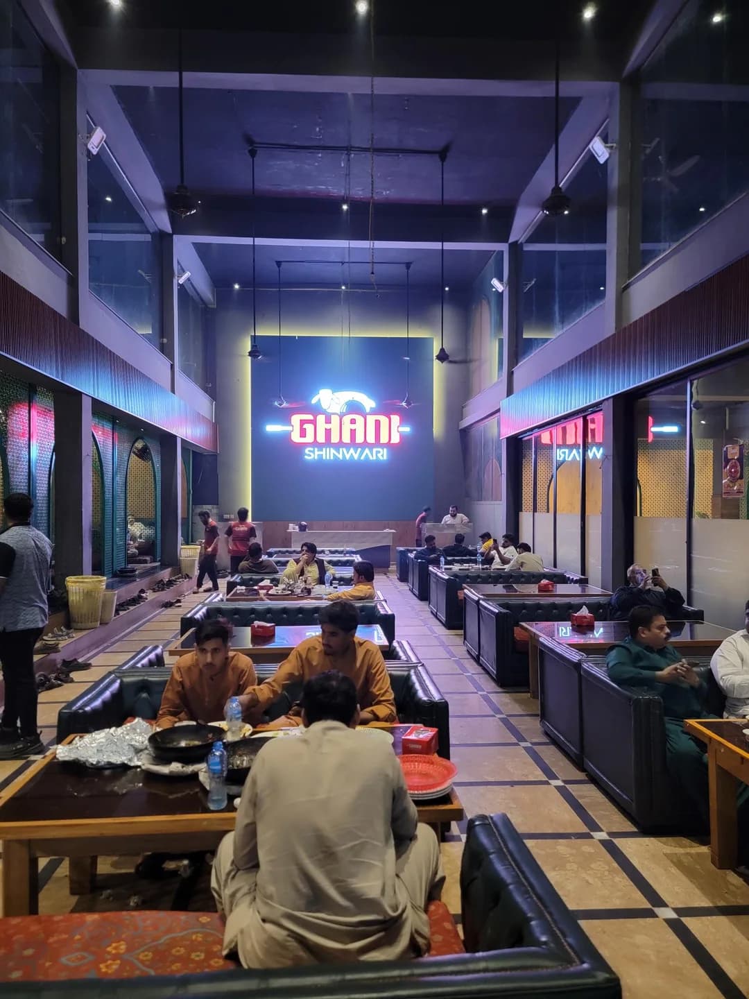 Guests dining beneath a high ceiling with the illuminated Ghani Shinwari sign at the end of the hall